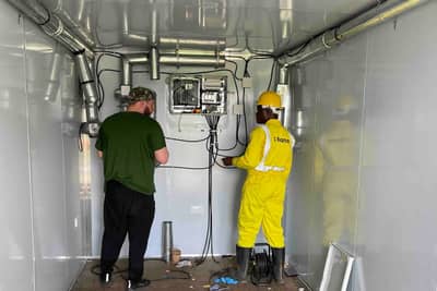 Two workers installing ventilation ducts and an electrical cabinet inside a facility module.